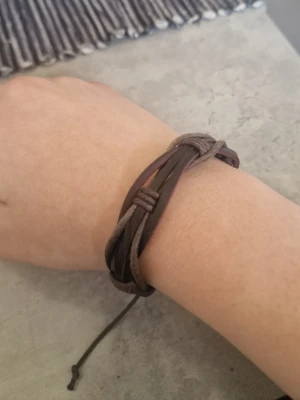 Brunt flätat läderarmband - Snyggt armband i brunt läder med flätad design och justerbar knytning. Armbandet har flera lager av tunna och bredare remmar som ger en cool och avslappnad look. Perfekt accessoar för dig som gillar en naturlig och stilren stil.