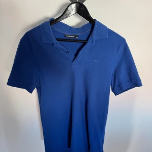 Blå pikétröja från J.Lindeberg - Snygg blå pikétröja från J.Lindeberg med klassisk krage och korta ärmar. Tröjan har två knappar vid halsen och diskret logga på bröstet. Tillverkad i mjukt bomullsmaterial som känns bekvämt hela dagen.