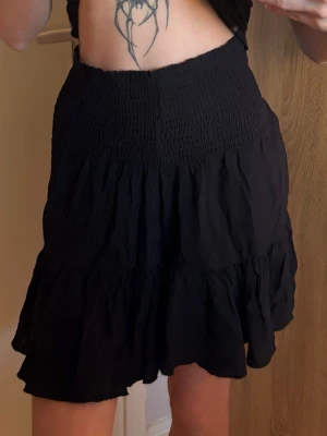 Svart volangkjol med smock - Supersöt svart kortkjol med smock i midjan och volang nertill. Kjolen har en luftig och flowy känsla, perfekt för varma dagar. Enkel att matcha med olika toppar för en trendig look.