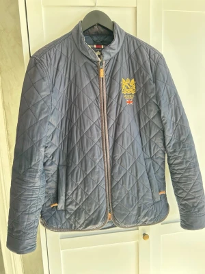 Mörkblå quiltad jacka från Morris - Snygg mörkblå quiltad jacka från Morris med gul broderad logga och brittisk flagga på bröstet. Jackan har dragkedja framtill, ståkrage och diskreta fickor. Perfekt för dig som gillar klassisk och stilren design.