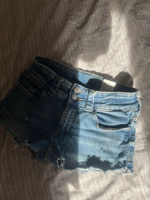 Blå jeansshorts med rå kant - Snygga blå jeansshorts med råa kanter och slitna detaljer. Klassisk femficksmodell med bälteshällor och dubbla knappar framtill. Perfekta för varma dagar och enkel att matcha med allt.