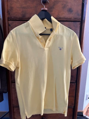 Gul pikétröja från Gant - Snygg gul pikétröja från Gant med klassisk krage och korta ärmar. Tröjan har knappar vid halsen och en liten broderad logga på bröstet. Tillverkad i mjuk bomull som känns skön mot huden. Perfekt för en avslappnad och stilren look.