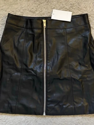 Svart kort skinnkjol med dragkedja - Snygg svart kort kjol i skinnimitation med en markerad silverfärgad dragkedja framtill. Kjolen har en rak passform och stilrena sömdetaljer både fram och bak. Perfekt att matcha med en tuff jacka eller stickad tröja.