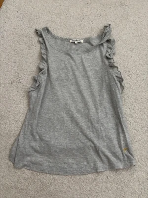 Grå linnetopp med volang från Hampton Republic - Snygg grå ärmlös linnetopp från Hampton Republic med volangdetaljer längs axlarna. Tillverkad i mjuk bomull som känns skön mot huden. Perfekt för en avslappnad och trendig look.