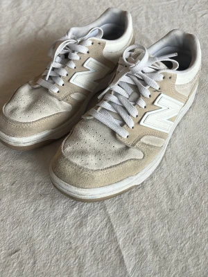 Beige New Balance sneakers i mocka - Snygga beige och vita sneakers från New Balance med klassisk N-logga på sidan. Ovandelen är i mocka och textil, med vita skosnören och perforerad tå för extra ventilation. Skorna har en låg silhuett och en bekväm sula som passar perfekt till streetwear.