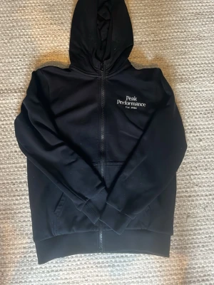 Svart Zip-hoodie från Peak Performance - Säljer en svart hoodie från Peak Performance med dragkedja och huva. Klassisk logga broderad i vitt på bröstet. Hoodien har fickor framtill och ribbade muddar. Perfekt för chill dagar eller när du vill ha en clean och enkel stil.