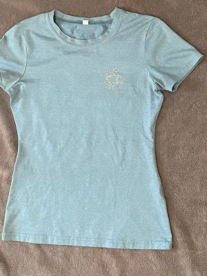 Rid T-shirt  - En ljusblå rid T-shirt som har en silver glitter detalj på den högra sidan. Skönt material att ha mot huden.