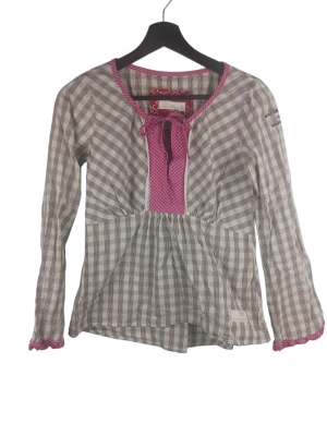 Odd Molly Wigwam rosa tröja. S - Rutig blus med långa ärmar och en v-ringning med knytband framtill. Blusen har en insydd resår i midjan som skapar en smickrande silhuett. Ärmsluten är utsmyckade med en virkad kant i en kontrasterande färg. På ena ärmen finns en broderad text. Flerfärgad tröja, kolla noggrant 🙂 Armhåla till armhåla: 41cm Axelbredd: 33cm Långt: 55cm Armlängden från armhåla: 44cm