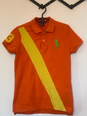 Orange Ralph Lauren t-shirt med tryck - Snygg orange t-shirt från Ralph Lauren med gul diagonal rand och stort grönt logotryck på bröstet. Nummer 3 i gult på ena ärmen och ryggen. Klassisk pikékrage och korta ärmar. Perfekt för dig som gillar sportig stil.