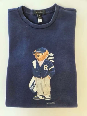 Mörkblå RLX Ralph Lauren sweatshirt polo bear  - Mörkblå sweatshirt från RLX Ralph Lauren med ikonisk björn i collegejacka och keps tryckt på framsidan. Tröjan har rund halsringning och är tillverkad i mjukt material. Perfekt för dig som gillar golf och snygga detaljer.