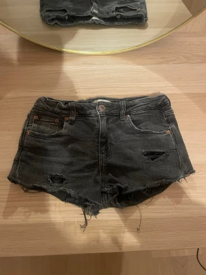 Svarta jeansshorts med slitningar - Säljer ett par svarta jeansshorts med råa kanter och slitna detaljer framtill. Klassisk femficksmodell med knapp och dragkedja. Perfekta för en avslappnad och trendig look.kommer från ett par jeans från Gina Tricot men har klippt de till shorts själv.skulle säga att de sitter som en xs/s.skriv för fler bilder, pris kan diskuteras!💗