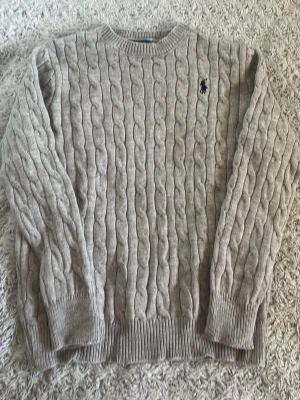 Grå kabelstickad tröja Polo Ralph Lauren - Kabelstickad tröja i ljusgrått från Polo Ralph Lauren. Klassisk rund halsringning, ribbade muddar och nederkant samt den ikoniska broderade loggan på bröstet. Tillverkad i mjukt material som ger en lyxig känsla.