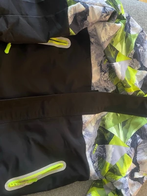 Färgstark vindjacka från Top&Sky Kids - Säljer en cool vindjacka från Top&Sky Kids med svart bas och färgglada detaljer i grönt, vitt och grått. Jackan har mönstrade partier, neongröna dragkedjor och en trekantig patch på ärmen. Perfekt för dig som gillar outdoor och streetstyle. Fleece på insidan som gör den perfekt för tidig vår eller höst jacka.