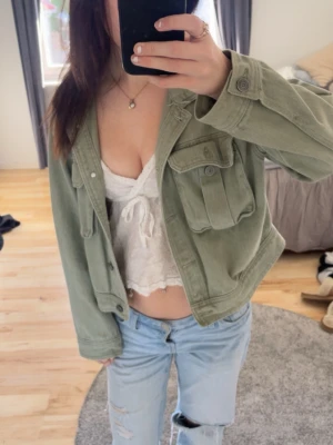 Grön jeansjacka med fickor - Snygg grön jeansjacka med två stora bröstfickor och knappar framtill. Jackan har en croppad passform och är perfekt att slänga över en topp eller hoodie. Stilren och enkel att matcha till olika outfits.