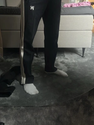 palm angels byxor ( nya modellen) - Säljer ett par svarta mjukisbyxor från Palm angels (nya modellen) med vita kontrast-ränder längs sidorna och dragkedja vid benslut. Byxorna har en avslappnad passform och broderad logga på vänster sida. Köpta på NK, nypris ca 5000