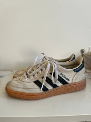 Adidas Spezial beige sneakers - Snygga Adidas Spezial sneakers i beige mocka med klassiska svarta ränder på sidorna och brun gummisula. Skorna har vita skosnören och låg profil, perfekt för en avslappnad streetwear-look. Diskret Adidas-logga på sidan och retrovibbar.