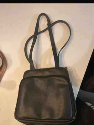 Svart läderhandväska med lång rem - En stilren svart handväska i läder med två långa axelband och en praktisk framficka med dragkedja. Väskan har en fyrkantig form och är perfekt för att bära dina viktigaste saker. Klassisk design som passar till många olika outfits.