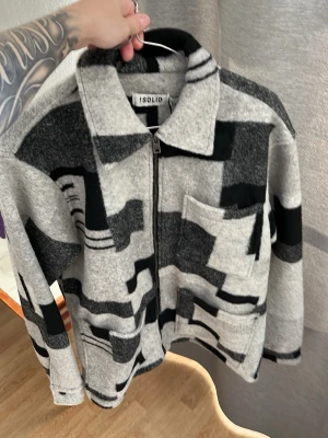 Gråsvart overshirt från Solid - Säljer en overshirt från Solid i grått och svart med grafiskt mönster. Jackan har dragkedja framtill, två stora fickor och en klassisk krage. Materialet ser ut att vara mjukt och fleece-liknande, perfekt för lager på lager. Overshirten har en avslappnad passform och modern look.