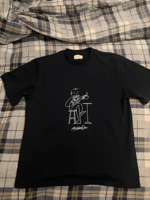 Mörkblå t-shirt från Aimé Leon Dore - Stilren och ikonisk T-shirt från Aimé Leon Dore med det klassiska “Café Reader”-motivet i vitt tryck. Motivet föreställer en avslappnad kille som sitter på en stol med en kopp ångande kaffe i handen och läser en tidning med “AIMÉ” på framsidan – ett av ALD:s mest tidlösa och populära grafiska motiv. •  Färg: Mörkblå (navy) •  Storlek: Medium (M) •  Skick: Nytt / oanvänt (aldrig buren, fortfarande med etiketter) •  Material: 100% bomull, skönt och mjukt •  Tillverkad i: Canada (datum på etiketten