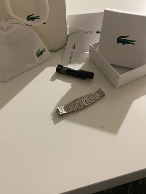Lacoste silverfärgat klockarmband - Säljer ett stilrent silverfärgat klockarmband från Lacoste. Armbandet är i metall och har en klassisk länkdesign. Medföljer originalförpackning, verktyg och påse med Lacoste-logga. Perfekt för dig som vill byta ut eller uppgradera din klocka.