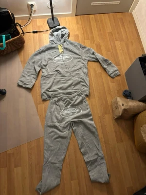 Corteiz set  - Mysigt grått loungewear-set från Café med hoodie och matchande byxor. Båda delar har ett vitt tryck med motiv på bröst och ben. Hoodien har känguruficka och dragsko i huvan. Materialet är mjukt och bekvämt, perfekt för chill hemma.