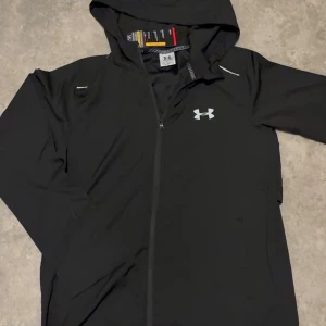 Svart hoodie från Under Armour - Snygg svart hoodie från Under Armour med dragkedja framtill och huva. Logga på bröstet och stort UNDER ARMOUR-tryck på ryggen. Tillverkad i ett lätt och sportigt material, perfekt för träning eller chill. Klassisk passform och stilren design.