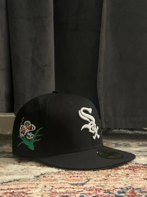 Svart White Sox keps med broderade fjärilar - Säljer en svart snapback keps från New Era med White Sox-logga framtill och broderade fjärilar i flera färger på sidorna. Kepsen har platt skärm och klassisk passform. Perfekt för dig som vill sticka ut med coola detaljer.