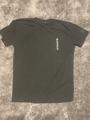 Svart Moncler t-shirt i bomull - Snygg svart t-shirt från Moncler med diskret logga tryckt på bröstet och en patch på ärmen. Klassisk passform och rund halsringning. Tillverkad i mjuk bomull som känns skön mot huden. Perfekt till jeans eller shorts.