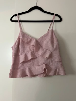 Ljusrosa volang topp med smala band - Supersöt ljusrosa topp med smala axelband och volangdetaljer. Toppen har en omlottkänsla och är tillverkad i ett lätt, strukturerat tyg som känns luftigt. Perfekt för varma dagar eller när du vill ha en romantisk vibe. Toppen har en dragkedja på sidan.