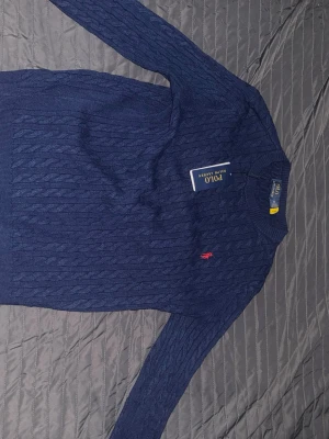 Marinblå kabelstickad tröja Polo Ralph Lauren - Kabelstickad marinblå tröja från Polo Ralph Lauren med klassisk röd broderad logga på bröstet. Tröjan har rund halsringning och långa ärmar. Perfekt för dig som gillar tidlös och stilren design.