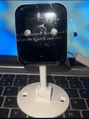 Vit övervakningskamera med stativ - Säljer en vit övervakningskamera med fyrkantig form och rundad front. Kameran har två synliga linser på framsidan och står på ett vitt stativ med monteringshål. USB-kabel för strömanslutning ingår. Perfekt för att hålla koll på hemmet eller kontoret.