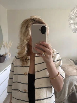 Randig beige overshirt med knappar - Snygg beige overshirt med svarta horisontella ränder och guldiga knappar framtill. Jackan har en boxig passform och trekvartsärm, perfekt att slänga över en topp för en trendig look.