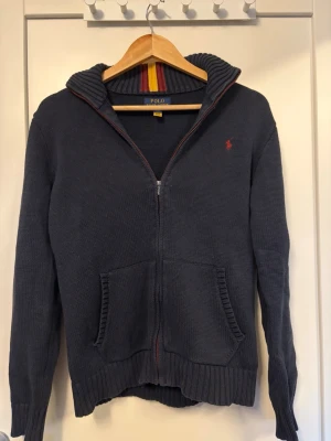 Mörkblå stickad hoodie från Polo Ralph Lauren - Mörkblå stickad hoodie med dragkedja från Polo Ralph Lauren. Tröjan har huva med ribbad kant, fickor framtill och röd logga broderad på bröstet. Insidan av kragen har ränder i rött och gult. Perfekt för en avslappnad och snygg stil.🔥😊🙌