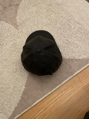 Svart keps från Prada i nylon - Säljer en svart keps från Prada med klassisk böjd skärm och justerbar rem baktill. Kepsen är tillverkad i nylon och har Pradas ikoniska metallogo på sidan. Perfekt för dig som vill ha en stilren och exklusiv accessoar.