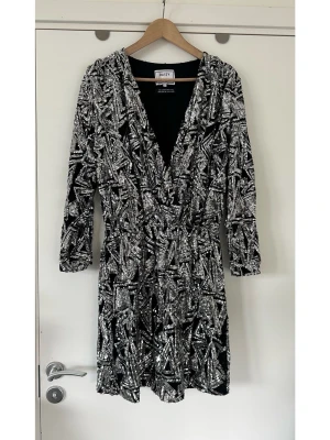 Glittrig paljett klänning från Daisy Street, storlek S - Underbar klänning från Daisy Grace i storlek S med iögonfallande design med paljetter som bildar ett abstrakt mönster över hela plagget. Klänningen har en djup V-ringning och långa ärmar med en knappdetalj vid ärmsluten. Midjan är markerad med resår, vilket ger en smickrande silhuett. Paljetterna är silverfärgade och kontrasterar effektfullt mot den svarta basen, vilket skapar en dynamisk och festlig look. Nypris 1600:-. Mycket gott skick, som ny!