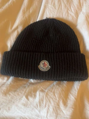 Svart Moncler mössa med logga - Snygg svart ribbstickad mössa från Moncler med uppvikt kant och klassisk Moncler-logga framtill. Perfekt accessoar för att hålla värmen och ge en stilren touch till din outfit.