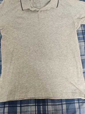 Ljusgrå pikétröja från Cubus - Snygg ljusgrå pikétröja från Cubus med klassisk krage och knappslå framtill. Tröjan har en diskret svart detalj på kragen och är tillverkad i mjukt bomullsmaterial. Perfekt för en clean och enkel stil.