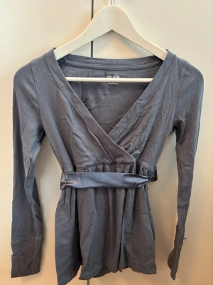 Blå omlottopp från Hollister - Snygg blå omlottopp från Hollister med lång ärm och v-ringad hals. Toppen har ett brett band i midjan som knyts för en markerad siluett. Materialet är ribbat och ger en skön känsla. Perfekt för dig som gillar stilrena och trendiga plagg.