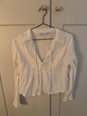 Vit blus med broderi från Zara - Säljer en vit blus från Zara med v-ringning och dekorativa broderier framtill. Blusen har långa ärmar, knytband vid halsen och vågig kant nedtill. Materialet är lätt och luftigt, perfekt för en trendig och avslappnad look.