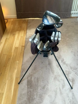 Golfset med bag och klubbor. Skriv för information om klubborna  - Golfset med svart golfbag och flera järnklubbor samt två drivers. Bagen har stödben och flera fack. Klubborna har svarta och röda detaljer, silverfärgade huvuden och gråa grepp. Perfekt för dig som vill ha ett komplett set till golfbanan.