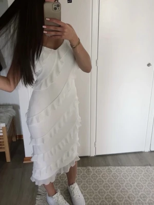 Vit klänning med volanger  - Säljer en snygg vit klänning med smala axelband och volangdetaljer i flera lager. Klänningen är knälång och har en figurnära passform. Perfekt för dig som gillar en stilren och trendig look.