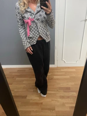 Mönstrad kofta med rosa rosett - Unik kofta med svartvitt nordiskt mönster och en iögonfallande rosa rosett på bröstet. Koftan har krage, långa ärmar och stängs framtill. Perfekt för dig som vill sticka ut med en cool detalj och gillar klassiska mönster med en twist.