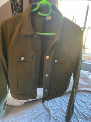 Olivgrön jeansjacka från H&M - Snygg olivgrön jeansjacka från H&M med klassisk krage, silverfärgade knappar och två bröstfickor med lock. Jackan har en rak passform och långa ärmar, perfekt att slänga över en hoodie eller t-shirt för en chill look.