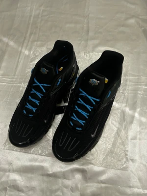 Nike Shox TL svarta/blå sneakers - Snygga Nike TN sneakers i svart mesh och syntet med blå detaljer och blå skosnören. Skorna har den ikoniska tn-sulan med fjädrande stötdämpning och små silverdetaljer. Perfekta för dig som gillar sportig och futuristisk stil.