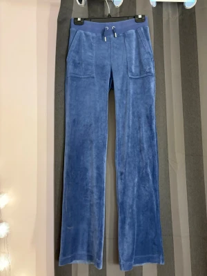 Blå lila velourbyxor Juicy Couture - Mjukisbyxor i blå velour från Juicy Couture med raka ben, bred resårmidja och dragsko. Byxorna har stora fickor fram och bakfickor med guldfärgade knappar med JC-logga. Perfekta för en chill och trendig look.
