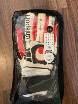 Uhlsport målvaktshandskar vit/röd - Säljer ett par Uhlsport målvaktshandskar i vitt, rött och svart med bred kardborrestängning. Handskarna har cool design och är perfekta för fotbollsträning eller match. Syntetmaterial för bra grepp och komfort.