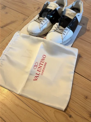 Valentino Garavani vita sneakers - Snygga vita sneakers från Valentino Garavani med bred svart lackdetalj över sidan. Klassisk låg modell med vita skosnören och rund tå. Kommer med dustbag och äkthetskort. Perfekta för dig som vill ha en lyxig touch till din outfit. Har även fixat till de lite.