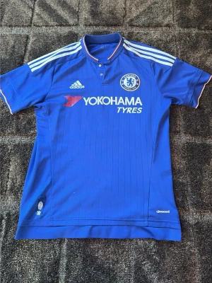 Chelsea FC matchtröja blå Adidas - Chelsea FC matchtröja i blått med vita detaljer och klubbmärke på bröstet. Tillverkad i lätt och ventilerande climacool-material. Adidas-logga och Yokohama Tyres sponsortryck på framsidan. Klassisk pikékrage med knappar och korta ärmar. Finns en defekt då sponsorn har flagnat bort lite där av priset. OBS ÄKTA 
