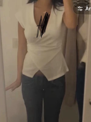 Vit omlottopp med v-ringning - Säljer en snygg vit omlottopp med v-ringning och korta ärmar. Toppen har en figurnära passform och en unik omlottdetalj nedtill som ger en modern look. Perfekt att styla med jeans eller kjol för en trendig outfit.