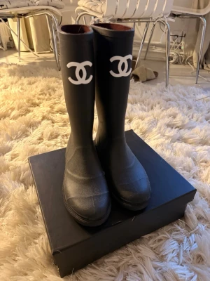 Chanel boots - Säljer ett par svarta höga gummistövlar från Chanel med den ikoniska vita CC-loggan framtill på skaftet. Stövlarna har en klassisk, stilren form och är perfekta för regniga dagar. Tillverkade i slitstarkt gummimaterial och har en rund tå.